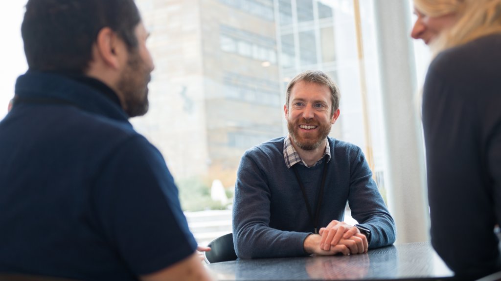 Smiling man sitting by a table while talking to two colleagues