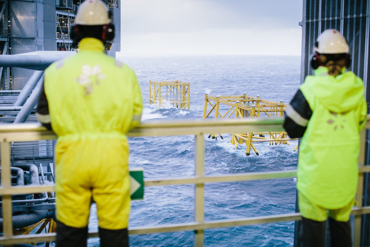 Photo of two people in protective gear standing at a railing and looking out on oil rig installations while offshore
