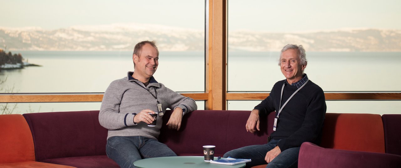 Two smiling men sitting in a sofa while drinking coffee and talking