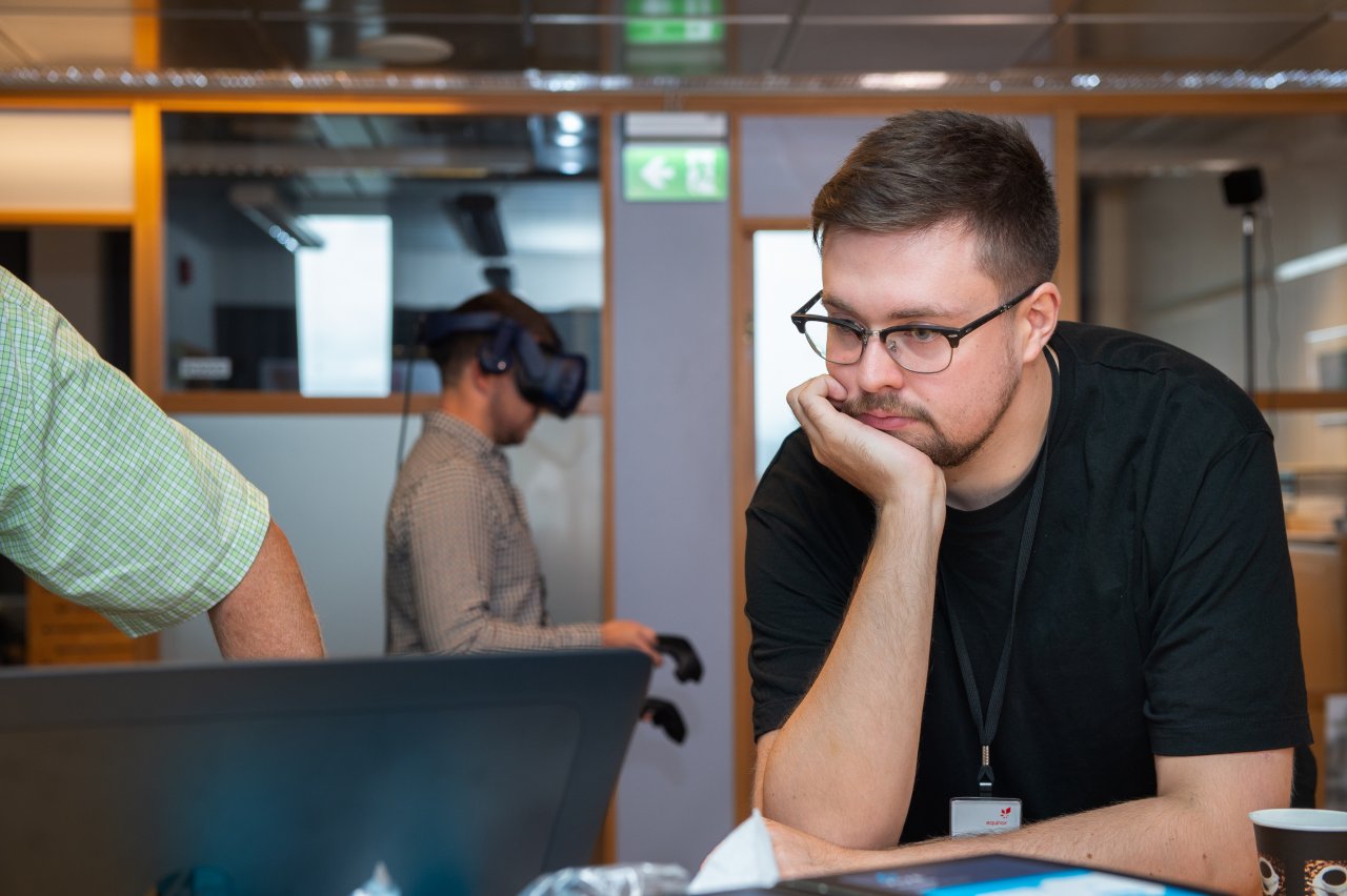 Man looking at a computer while another man wears a VR headset in the background